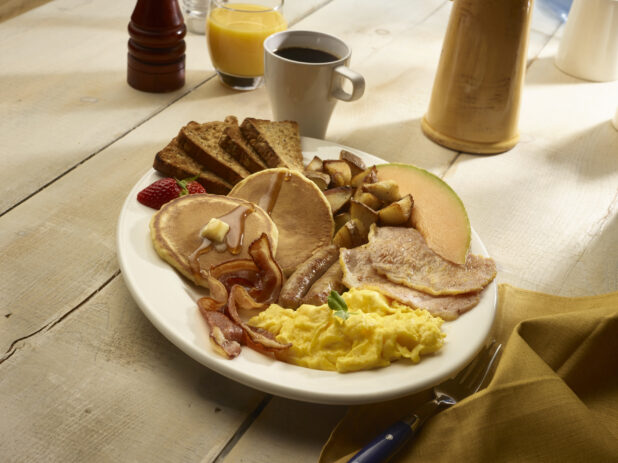 Big breakfast with scrambled eggs, canadian bacon, bacon, pancakes, home fries and toast with fruit on a rustic wooden table with accessories