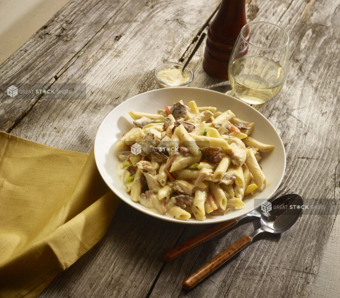 Chicken and mushroom penne pasta in a cream sauce in a white round bowl with a glass of white wine and parmesan cheese and accessories