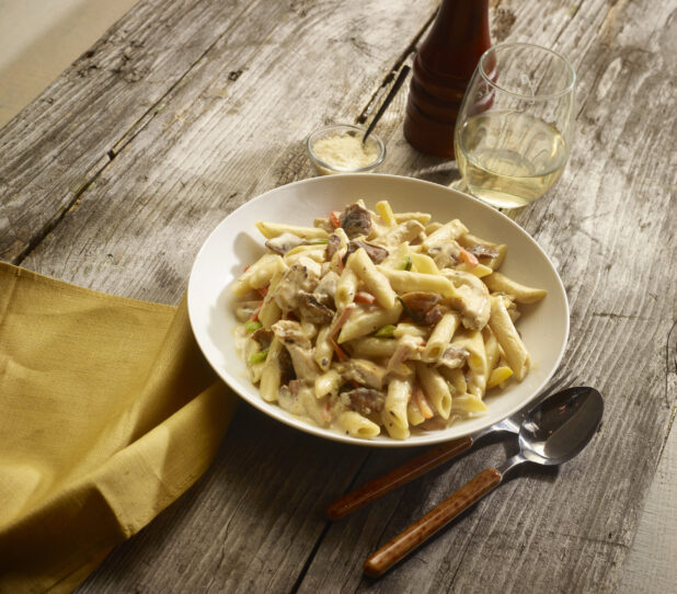 Chicken and mushroom penne pasta in a cream sauce in a white round bowl with a glass of white wine and parmesan cheese and accessories