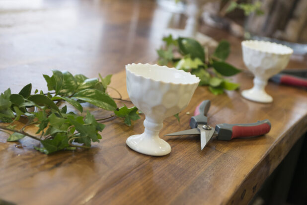 White flower goblet with greenery and ivy with shears in the foreground with a white flower goblet and greenery in the background on a wooden table