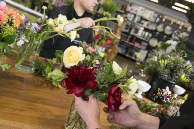 Hands arranging red peonies and yellow ranunculus in a glass vase on a wood work table