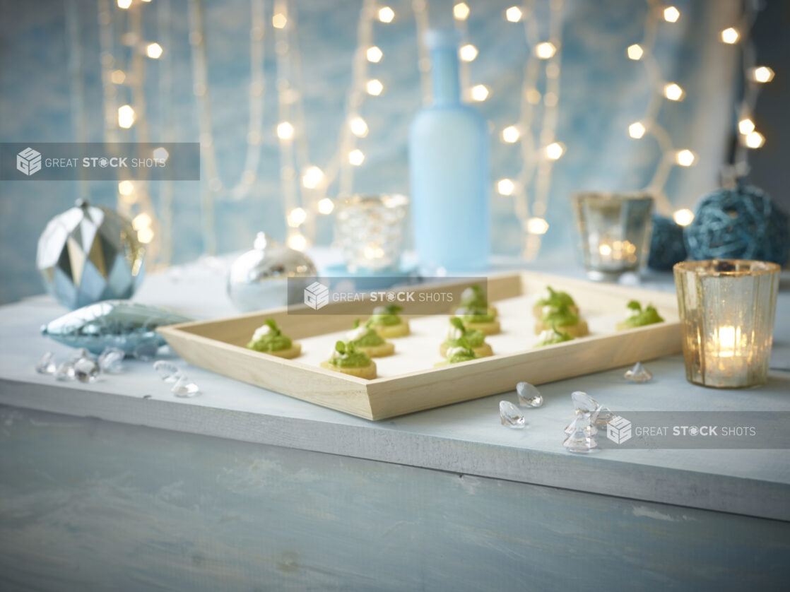 A Wood Serving Tray of Canapés with Pesto, Crème Fraiche and a Garnish of Micro Greens in a Blue Holiday Themed Indoor Setting with Tea Light Candles