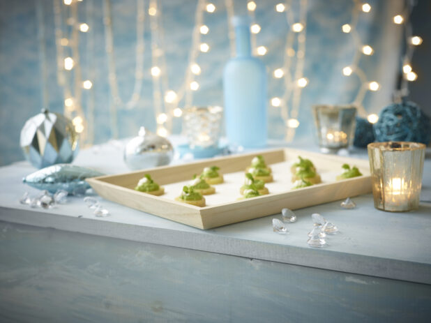 A Wood Serving Tray of Canapés with Pesto, Crème Fraiche and a Garnish of Micro Greens in a Blue Holiday Themed Indoor Setting with Tea Light Candles
