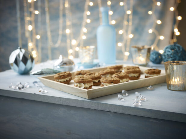 A Wood Serving Tray of Cream-Filled Cookie Sandwiches in a Blue Holiday Themed Indoor Setting with Tea Light Candles