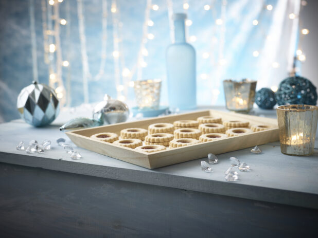 Jam filled Christmas cookies on a rectangular wood catering tray with ethereal light blue background with sparkling decorations