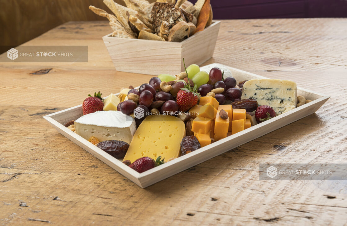 Cheese tray with various cheeses, nuts, fruits, dried fruit in a rectangular wood catering tray with assorted breads and crackers in a square wood catering box