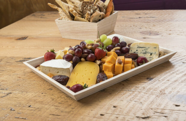 Cheese tray with various cheeses, nuts, fruits, dried fruit in a rectangular wood catering tray with assorted breads and crackers in a square wood catering box