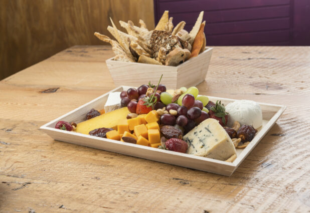 Assorted cheese and fruit platter on a wood catering tray with breadsticks in the background