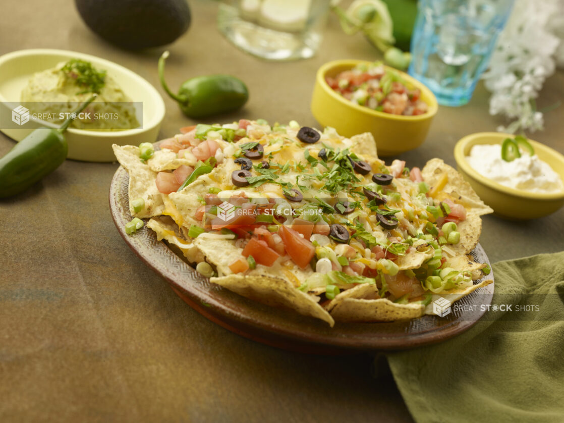 Fully loaded nachos on a round ceramic platter with guacamole, pico de gallo and sour cream with accessories in the background on a rustic background