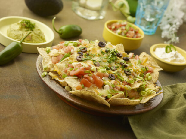 Fully loaded nachos on a round ceramic platter with guacamole, pico de gallo and sour cream with accessories in the background on a rustic background