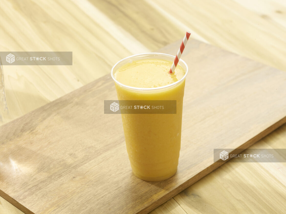 Orange smoothie in a plastic cup with a red and white straw on a wooden board on a wooden background