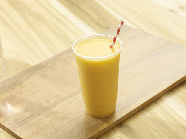 Orange smoothie in a plastic cup with a red and white straw on a wooden board on a wooden background