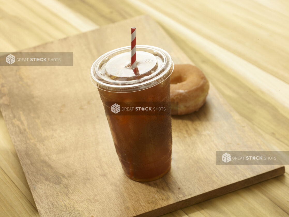 Iced tea in a clear plastic cup with a lid and red and white straw with a donut in the background on a wooden background