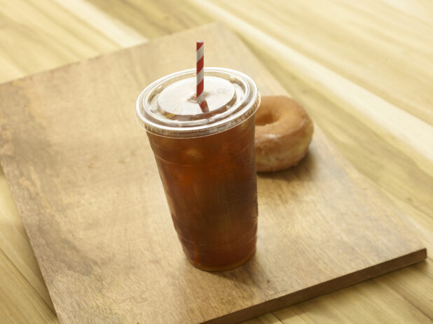 Iced tea in a clear plastic cup with a lid and red and white straw with a donut in the background on a wooden background