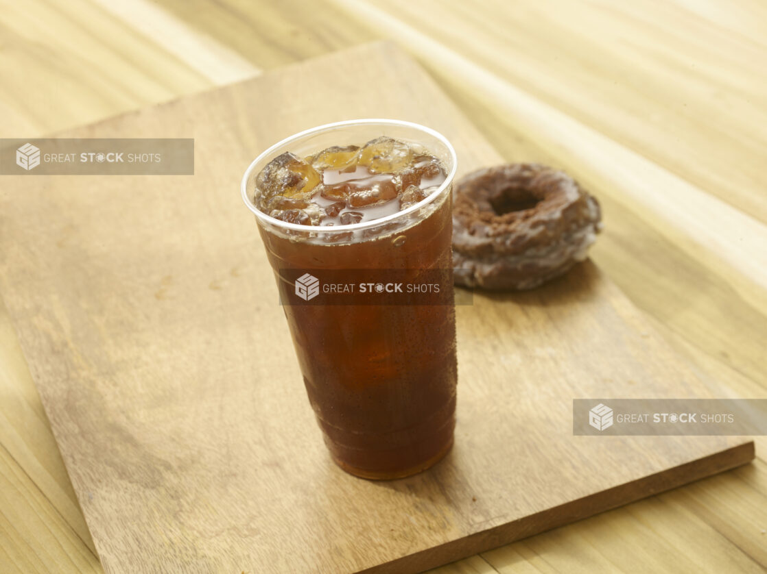 Iced tea in a clear plastic cup with a chocolate donut in the background on a wooden board on a wooden background