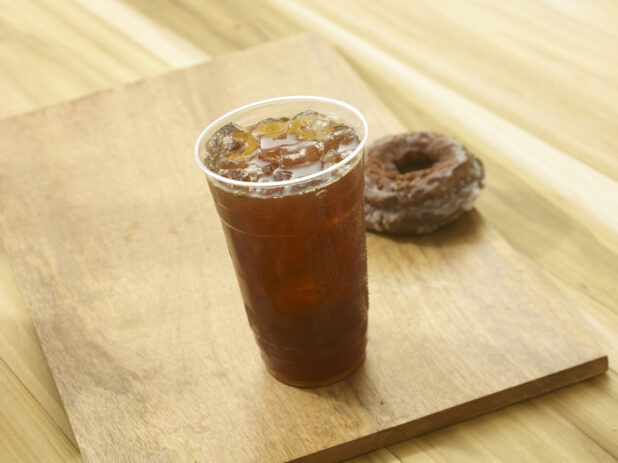 Iced tea in a clear plastic cup with a chocolate donut in the background on a wooden board on a wooden background