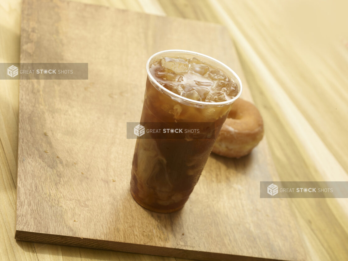 Iced coffee in a clear plastic cup with a donut on a wooden board on a wooden background