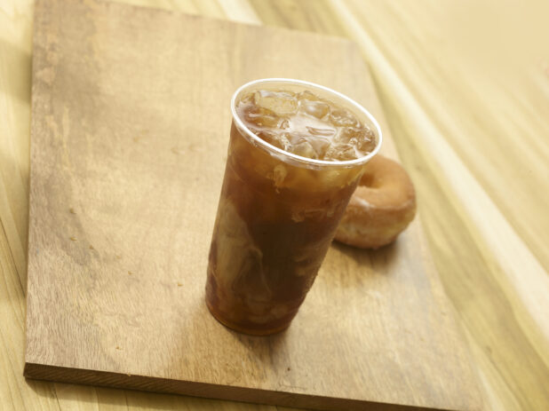 Iced coffee in a clear plastic cup with a donut on a wooden board on a wooden background