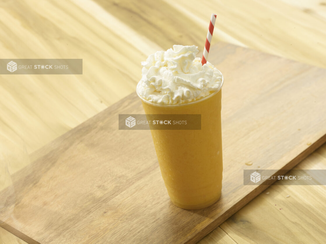 Mango/Orange fruit smoothie with whipped cream on top with a red and white striped straw on a wooden background