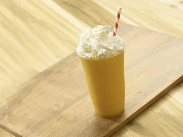 Mango/Orange fruit smoothie with whipped cream on top with a red and white striped straw on a wooden background