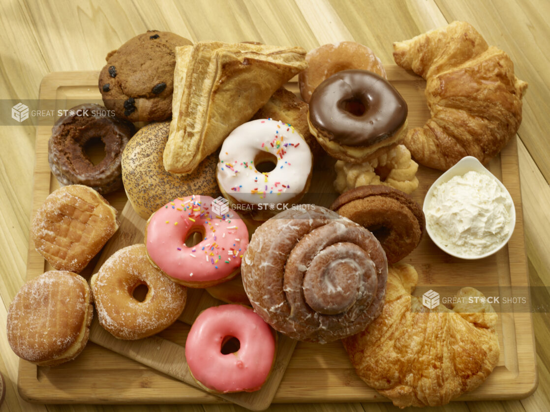 Assorted donuts, bagel, croissant, muffin and turnover on a wooden cutting board with a side of whipped butter on a wooden background