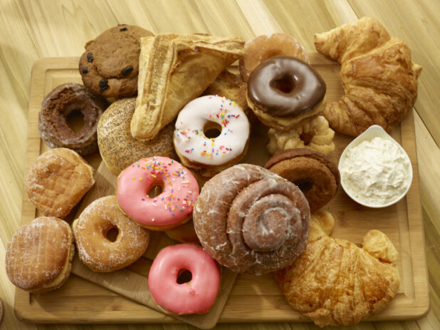 Assorted donuts, bagel, croissant, muffin and turnover on a wooden cutting board with a side of whipped butter on a wooden background