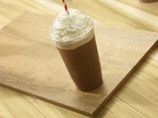 Iced coffee in a clear plastic cup with whipped cream, a clear dome lid and straw on a wooden board