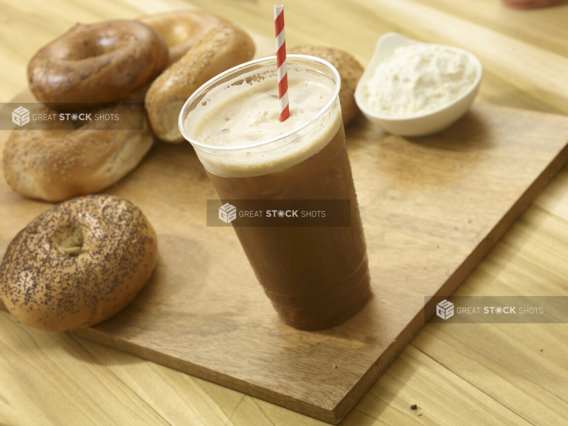 Iced coffee in a clear plastic cup with several whole bagels and a ramekin of cream cheese on a wooden board with a wooden background