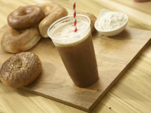 Iced coffee in a clear plastic cup with several whole bagels and a ramekin of cream cheese on a wooden board with a wooden background