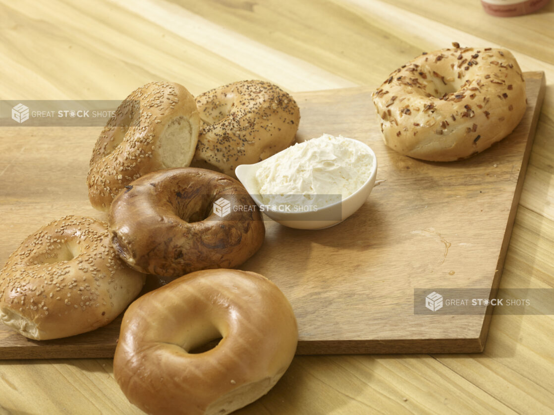 Various whole uncut bagels on a wooden board with a bowl of cream cheese on a wooden background