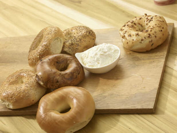 Various whole uncut bagels on a wooden board with a bowl of cream cheese on a wooden background