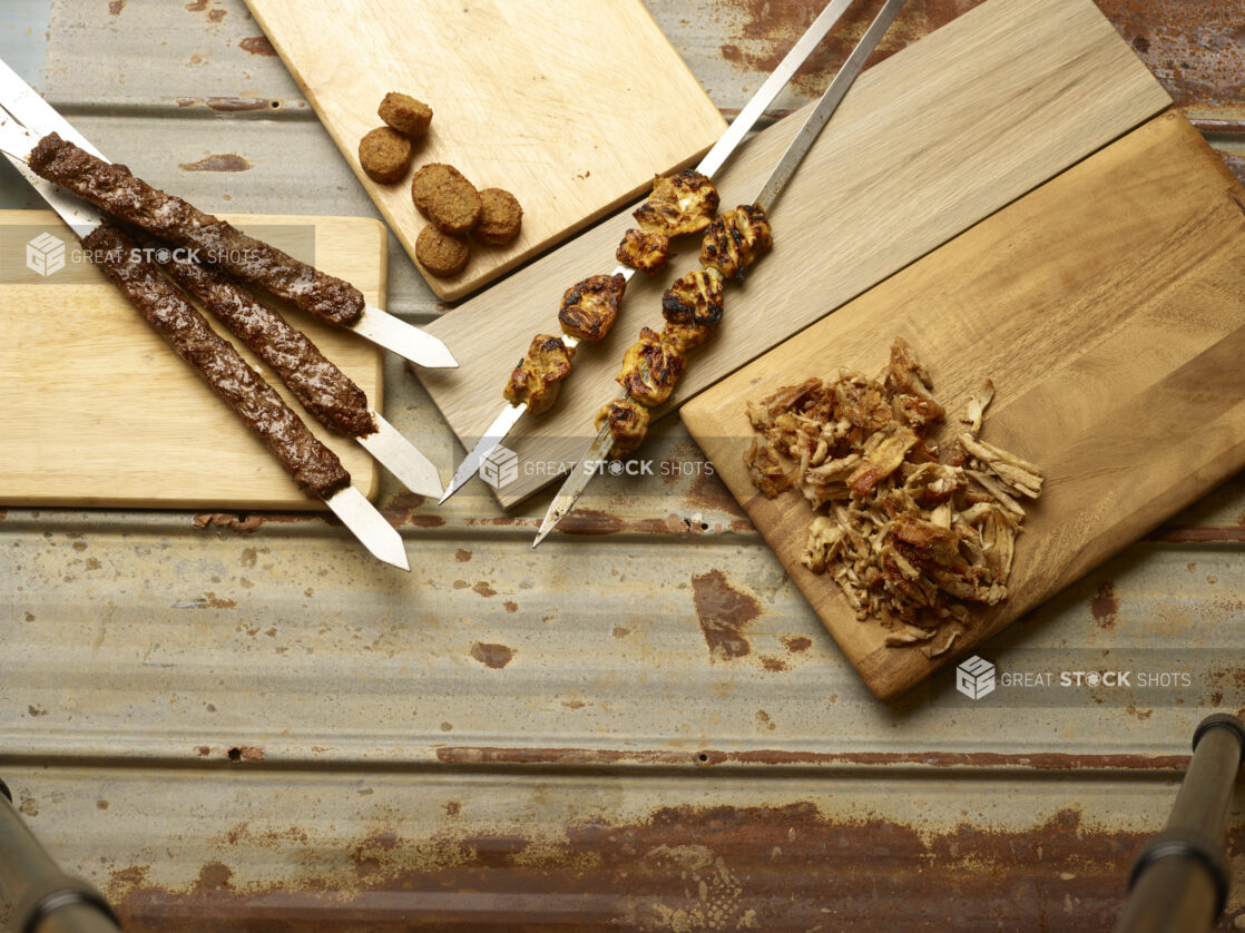 Middle Eastern meats and chicken on skewers, sliced chicken shawarma and falafel displayed on wooden boards on a rustic tin background