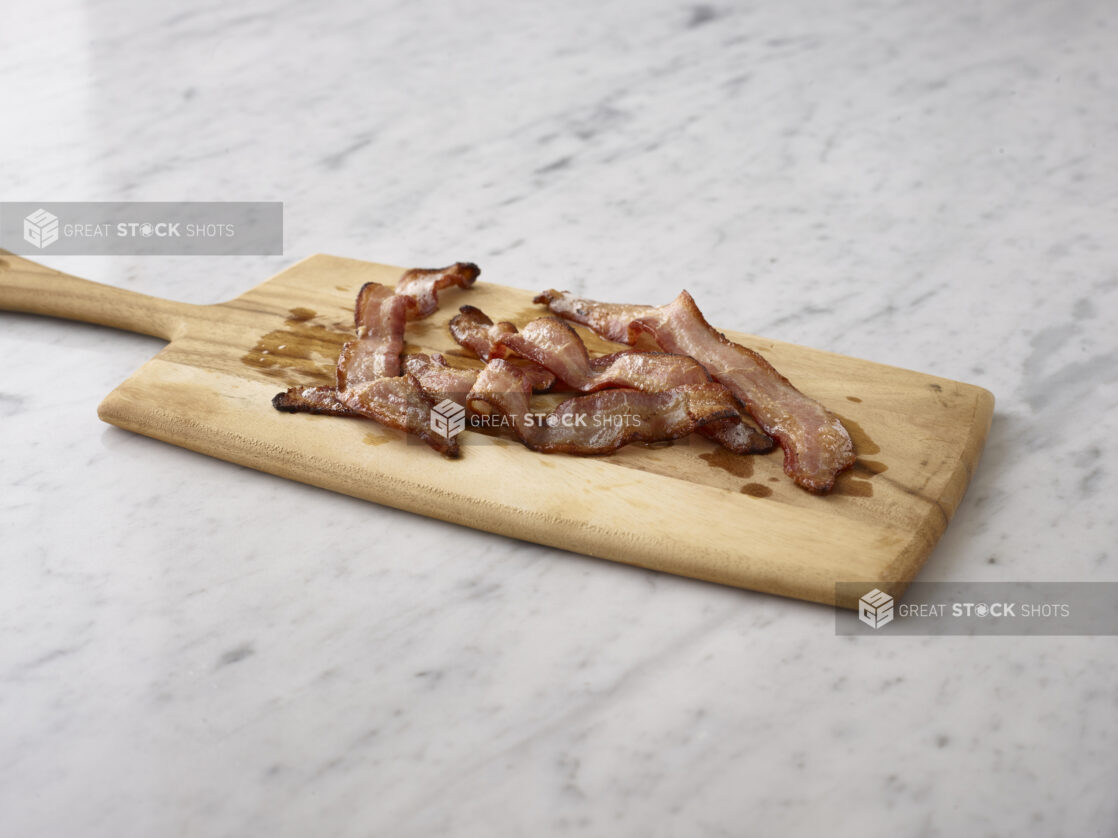 Slices of cooked bacon on a wooden cutting board on a white marble background