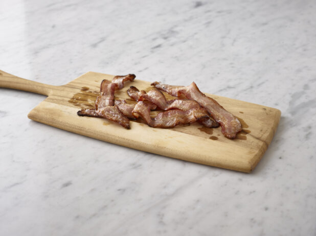 Slices of cooked bacon on a wooden cutting board on a white marble background