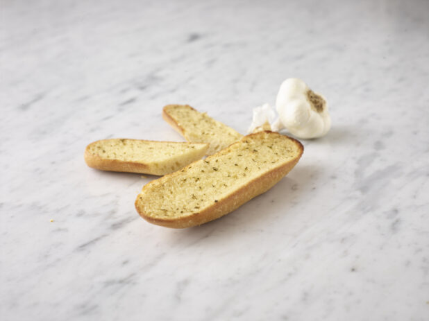 Garlic bread with a whole fresh garlic bulb all on a white marble background