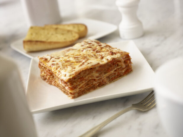 One piece of meat lasagna on a square white plate with a fork in the foreground and 2 pieces of garlic bread on a white rectangular plate in the background