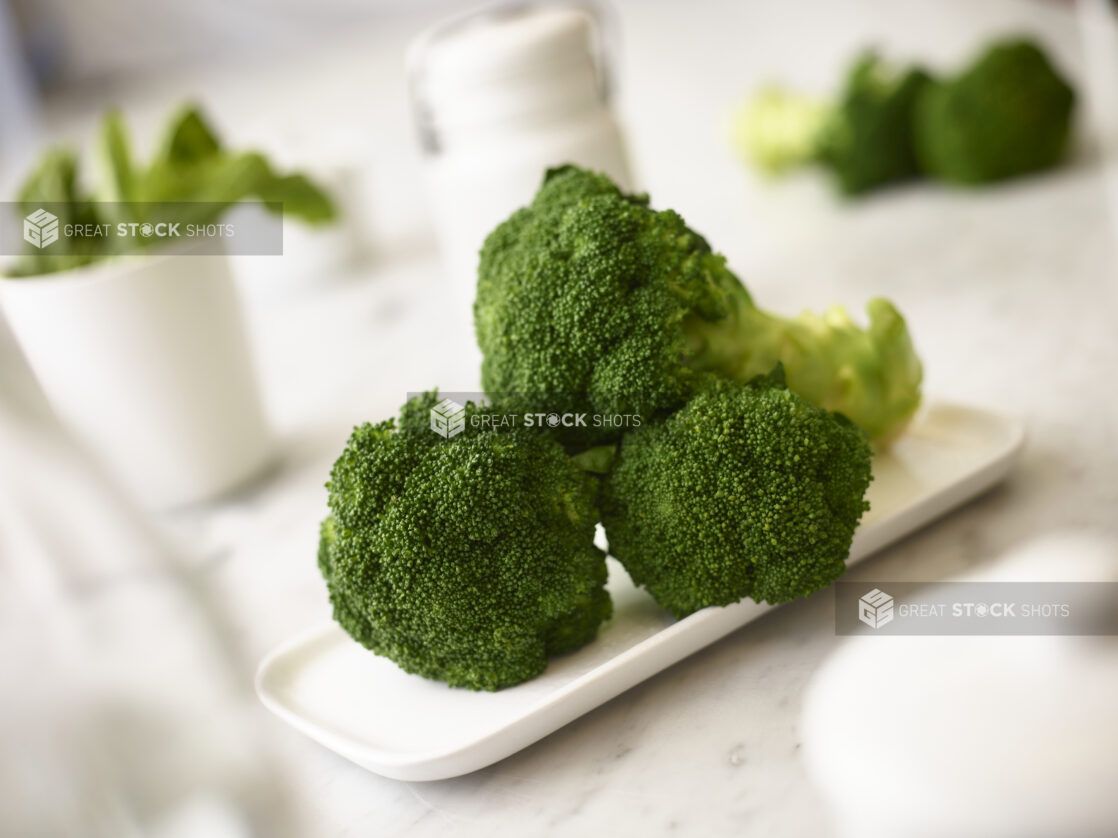 Fresh broccoli on a white rectangular plate on a white marble background with white accessories and fresh vegetables in the background