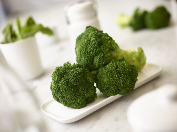 Fresh broccoli on a white rectangular plate on a white marble background with white accessories and fresh vegetables in the background