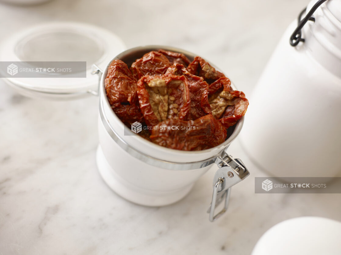 Sundried tomatoes in a white ceramic jar with additional white jars surrounding on a white marble background