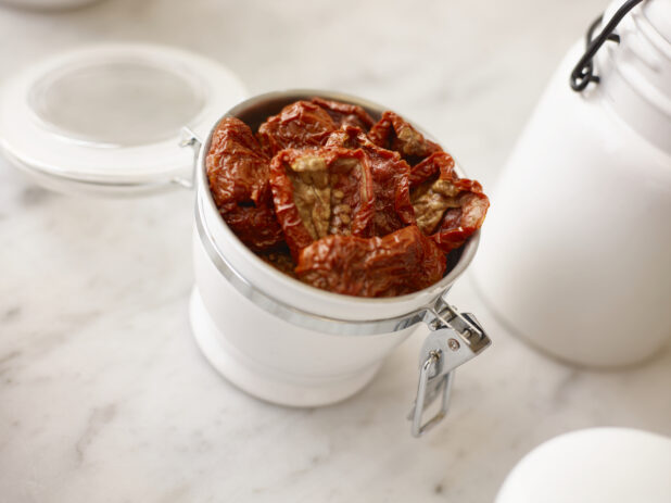 Sundried tomatoes in a white ceramic jar with additional white jars surrounding on a white marble background