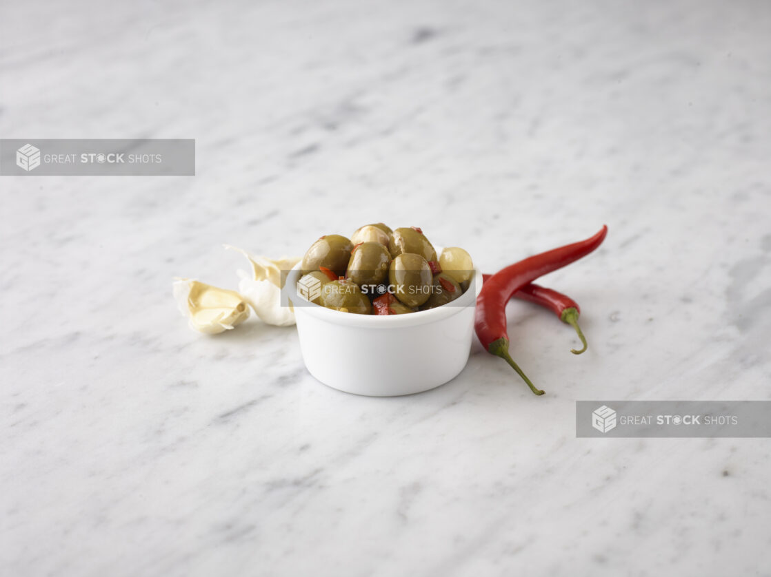 Marinated Queen olives in a round white ramekin with fresh red chili peppers and garlic cloves behind on a white marble background