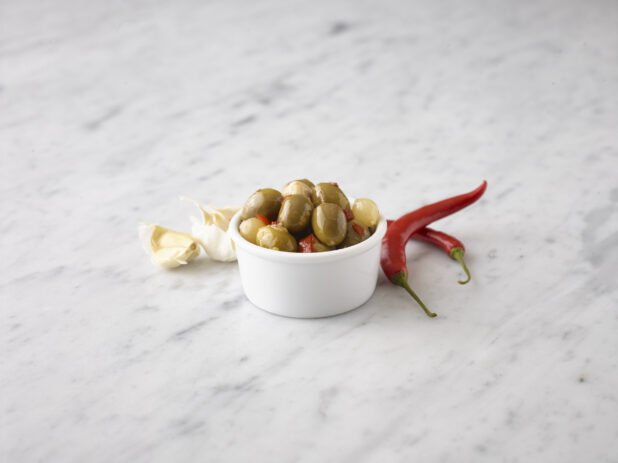 Marinated Queen olives in a round white ramekin with fresh red chili peppers and garlic cloves behind on a white marble background