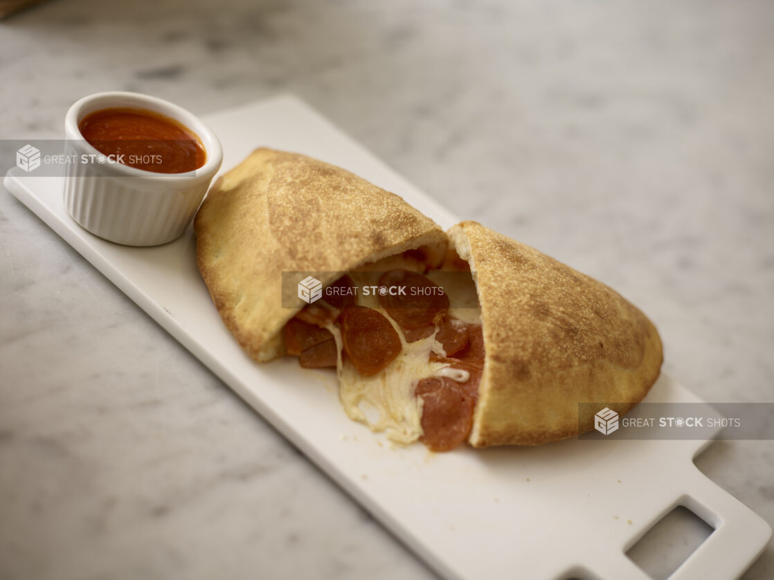 Pepperoni Calzone cut open with a round white ramekin of tomato sauce beside it on a white board on top of a white marble background