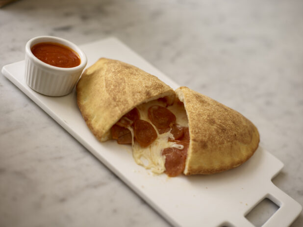 Pepperoni Calzone cut open with a round white ramekin of tomato sauce beside it on a white board on top of a white marble background