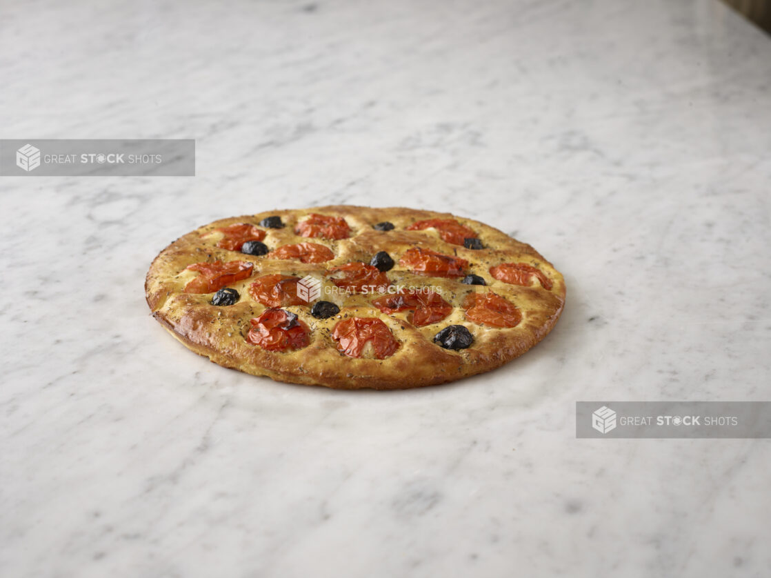 Round focaccia with black olives and roasted tomatoes on a white marble background