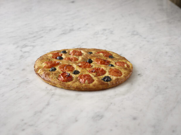 Round focaccia with black olives and roasted tomatoes on a white marble background