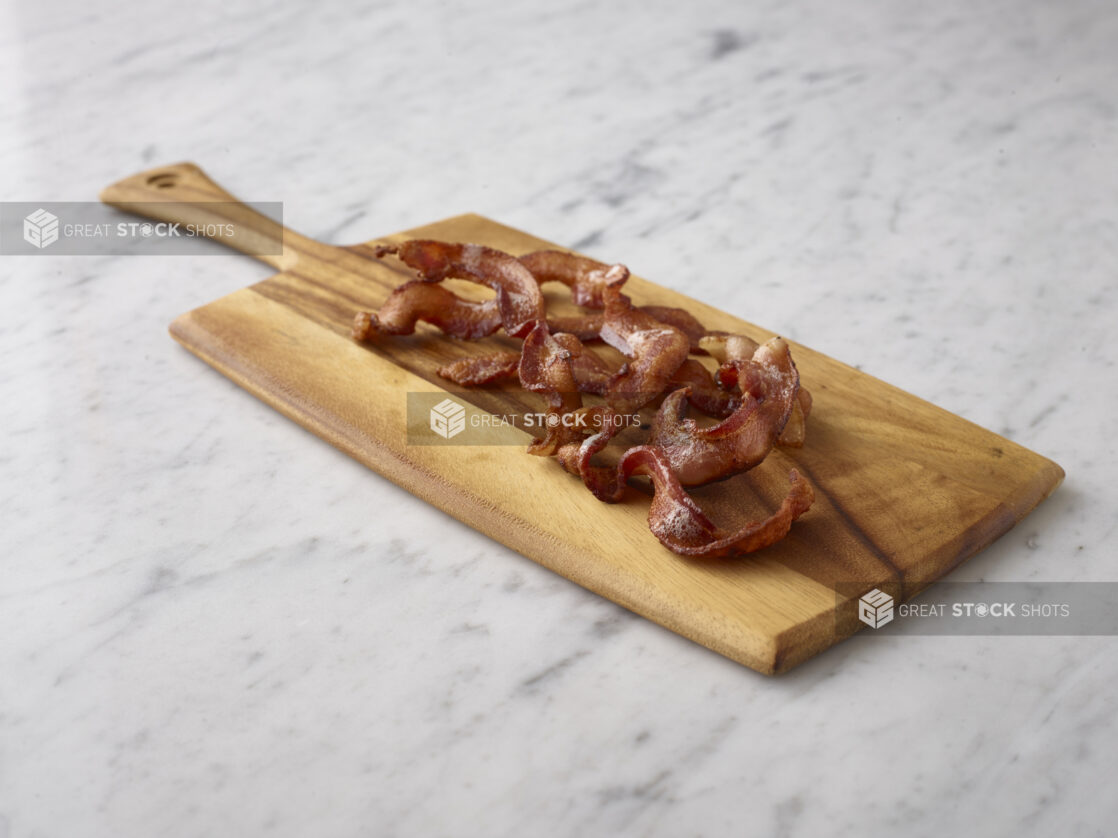 Sliced cooked bacon on a wooden cutting board on a white marble background