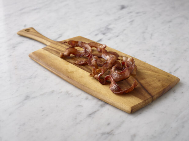 Sliced cooked bacon on a wooden cutting board on a white marble background