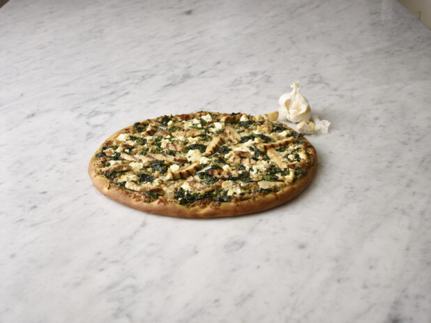 Whole 3 topping pizza with chicken, feta cheese and spinach on a white marble background with a garlic bulb and cloves in the background