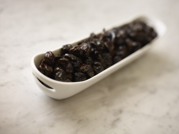 Whole black pitted olives in a long cylindrical white dish on a white marble background